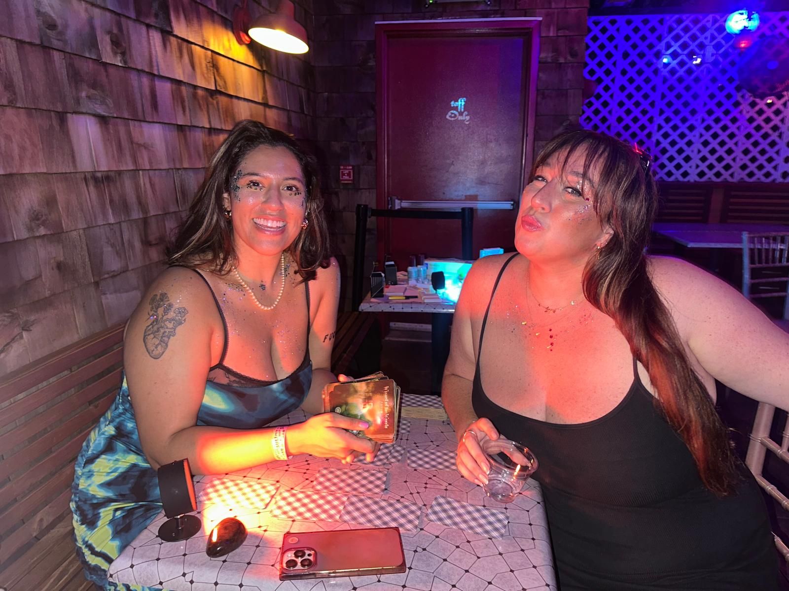 Kaide and a guest smiling at a bar table with a tarot deck, lit by a small lamp